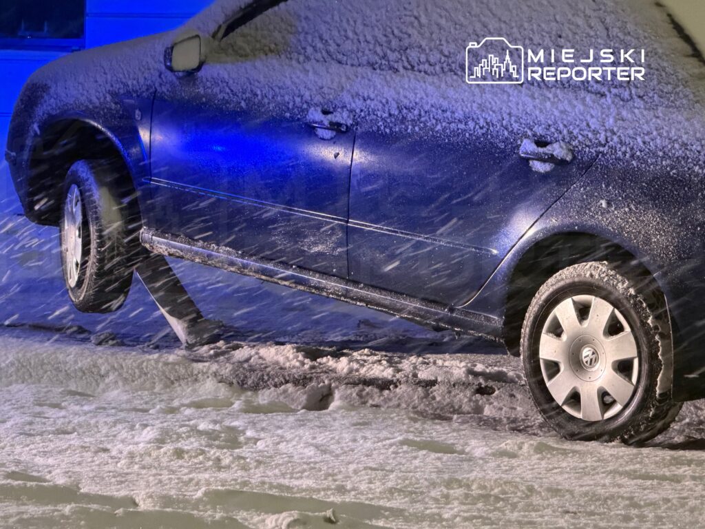 Niebieski samochód unosi się na śniegu, z jednym kołem w powietrzu, w intensywnym opadzie śniegu.