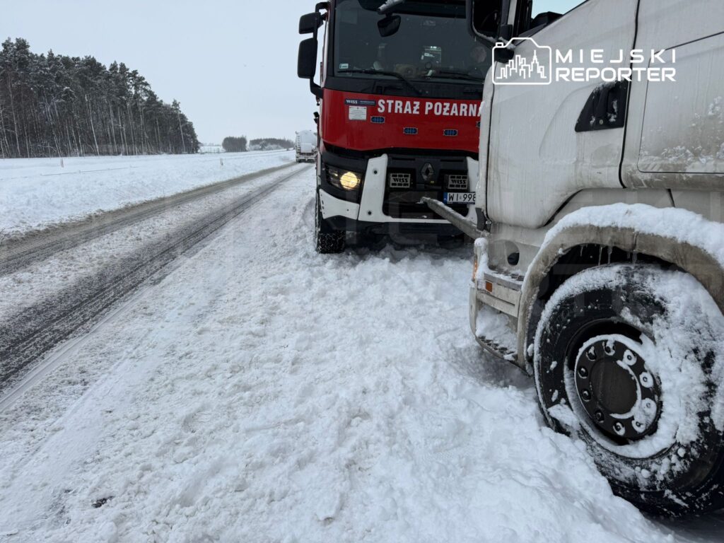 Czerwony wóz strażacki stoi na zaśnieżonej drodze, obok białego ciężarowego pojazdu utknęłego w śniegu.