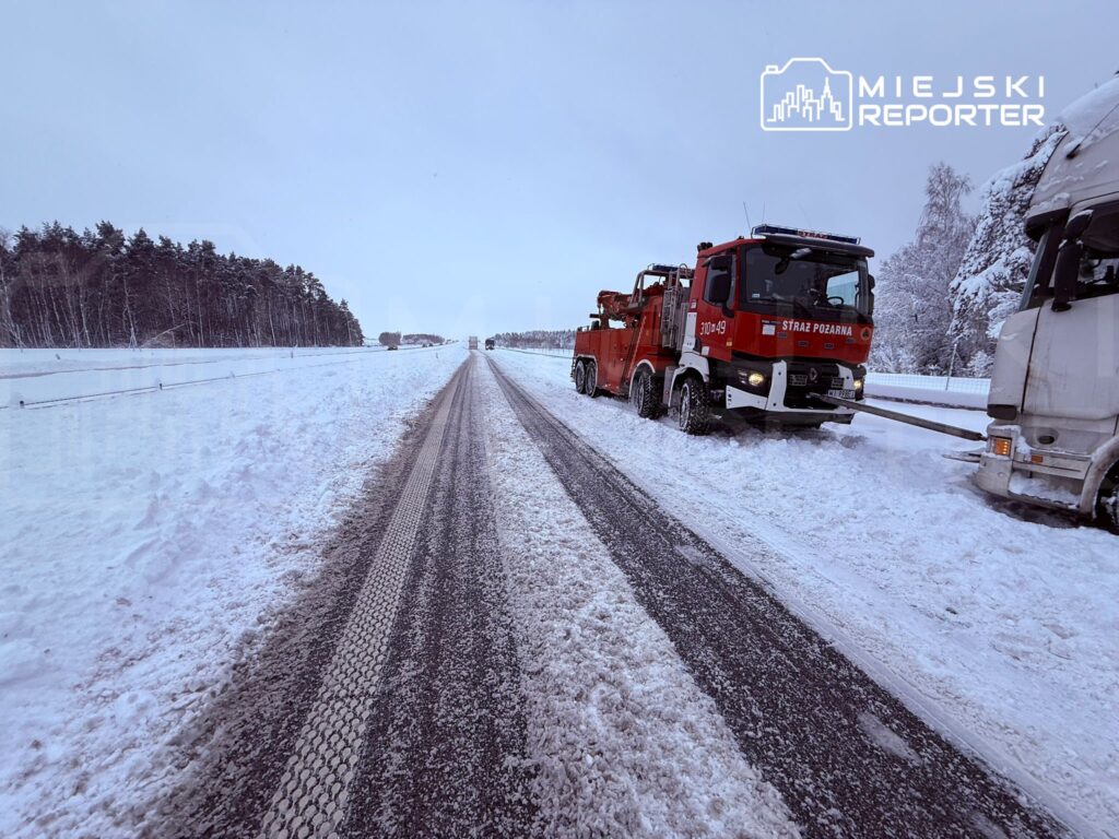 Czerwony wóz straży pożarnej holuje ciężarówkę na zaśnieżonej drodze, otoczonej lasem.