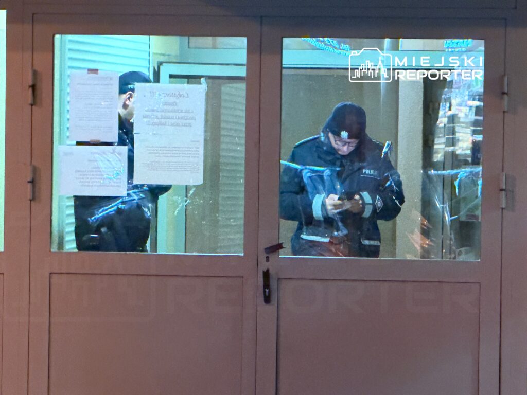 Policjant w mundurze przegląda telefon komórkowy, stojąc za przeszklonym wejściem budynku.