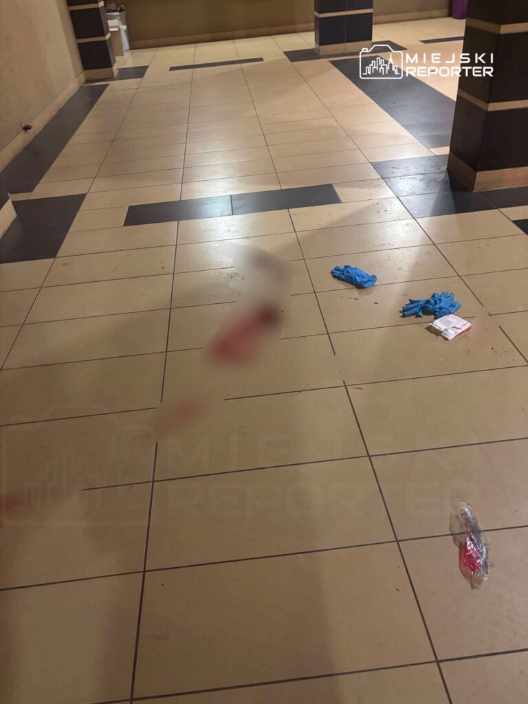 Na posadzce w korytarzu widoczne są plamy krwi oraz niebieskie rękawiczki. Obok leży zapakowany przedmiot.