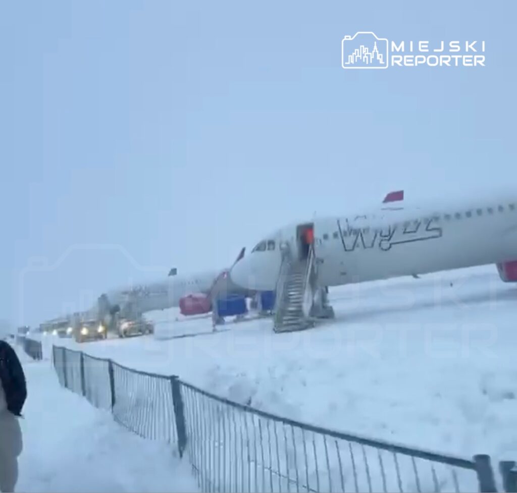 Samoloty na lotnisku otoczone śniegiem, z pracownikami przy schodach wyjściowych w złych warunkach pogodowych.