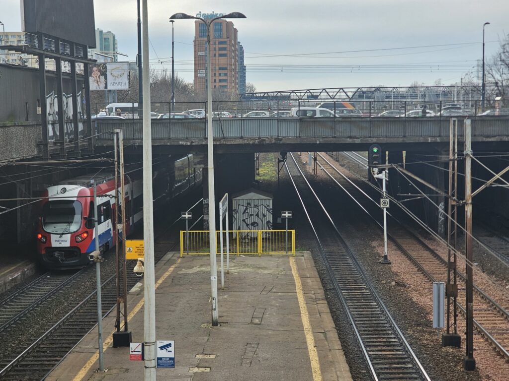 Pociąg w kolorze czerwono-niebieskim wjeżdża na stację kolejową, pod przeszklonym wiaduktem, obok torów.