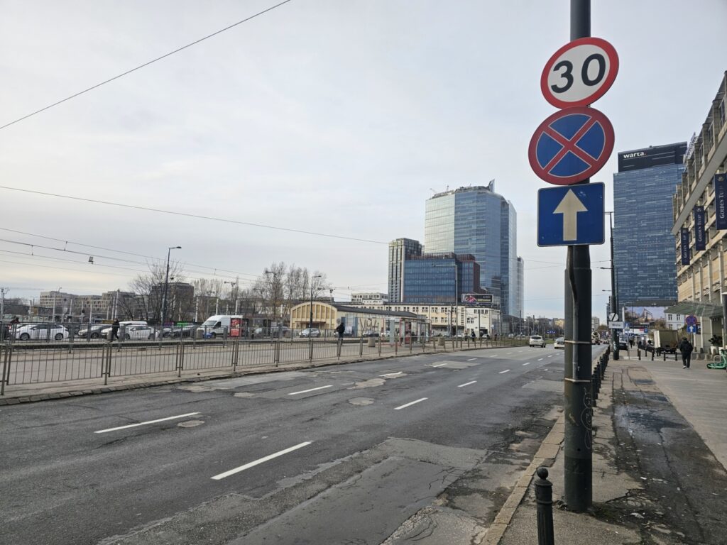 Ulica z widokiem na nowoczesne biurowce, znaki drogowe ograniczające prędkość do 30 km/h oraz zakaz parkowania.