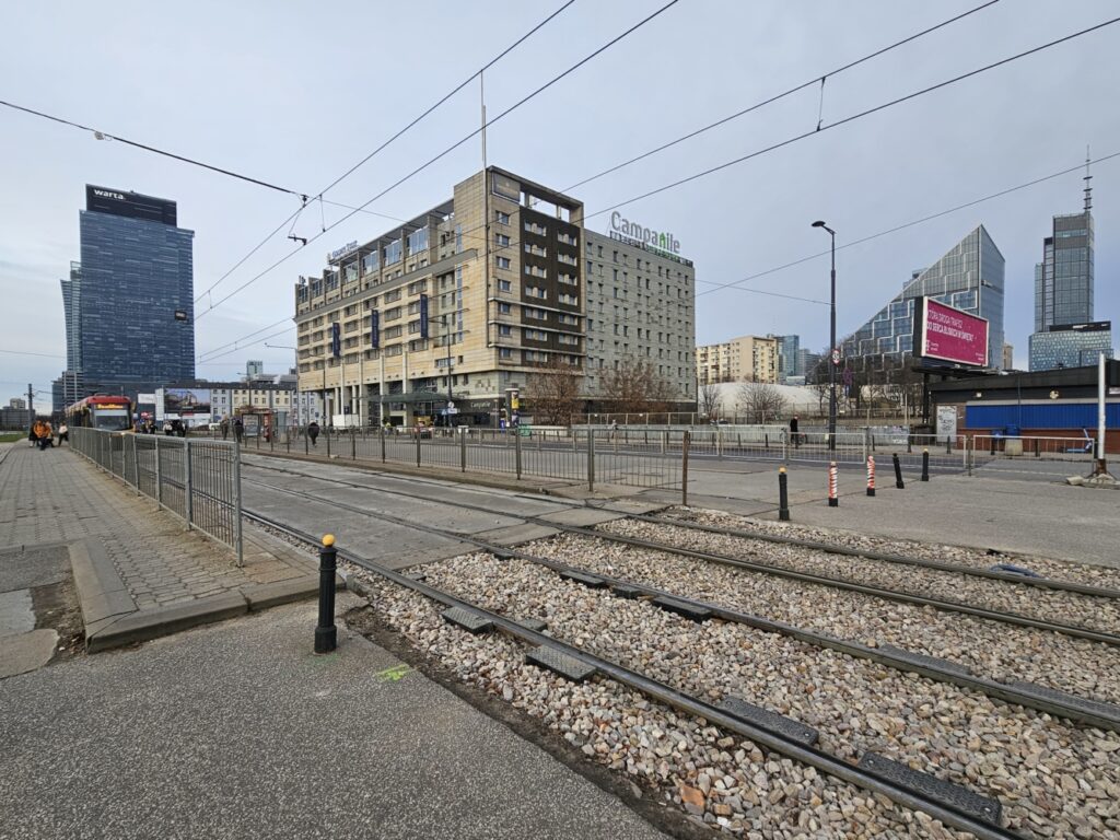 Nowoczesne biurowce w tle, tramwaje na torach, piesi na chodniku przy ruchliwej ulicy w miejskim otoczeniu.