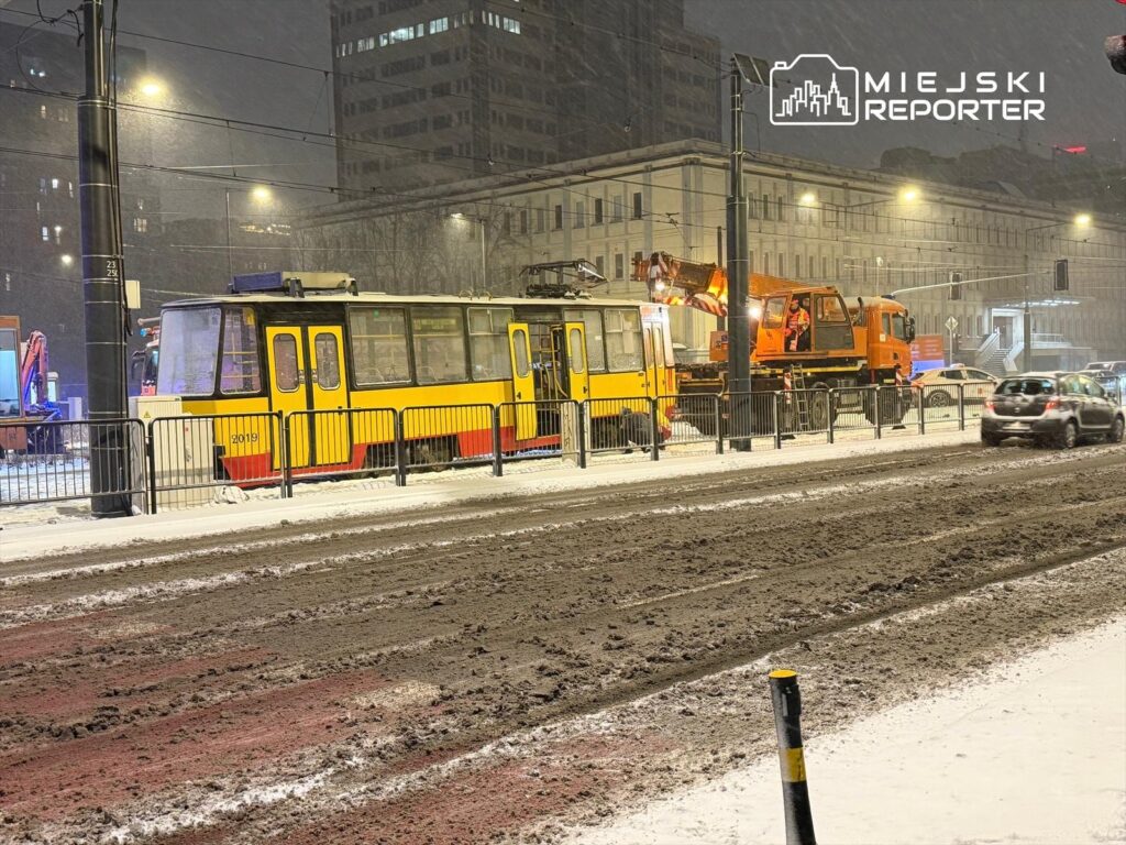 Żółty tramwaj stoi na zaśnieżonej ulicy, obok niego pracuje pomarańczowa maszyna do odśnieżania.