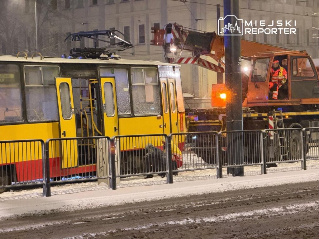 Tramwaj z żółtą i czerwoną powłoką stoi na zaśnieżonej ulicy, obok pracownik w odblaskowej kamizelce obsługuje dźwig.