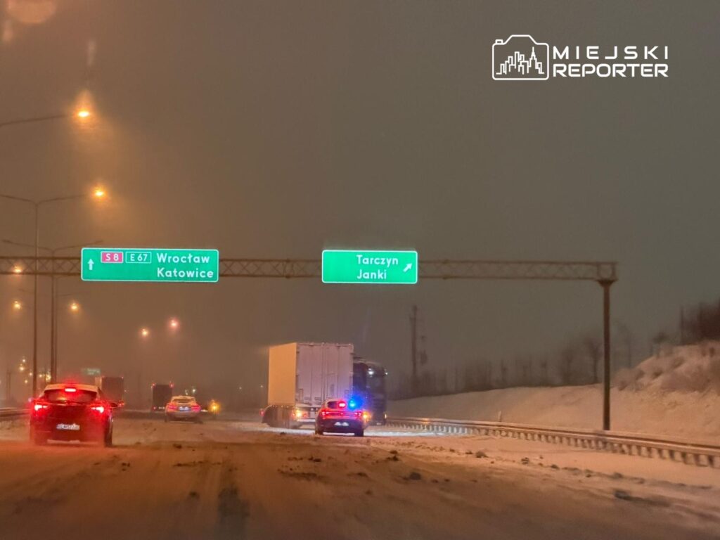 Na zaśnieżonej drodze widać samochody osobowe oraz ciężarówkę, a nad nimi wiszą znaki drogowe wskazujące kierunki.