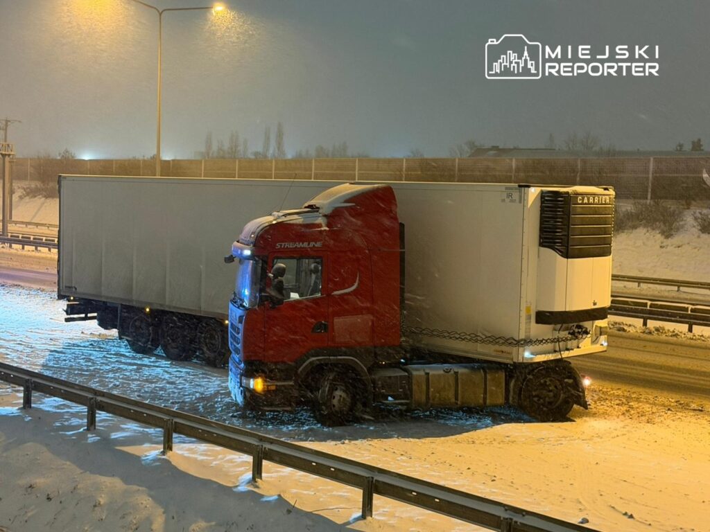 Czerwony ciężarowy pojazd z naczepą stoi na zaśnieżonej drodze w nocy, otoczony przez opadający śnieg.
