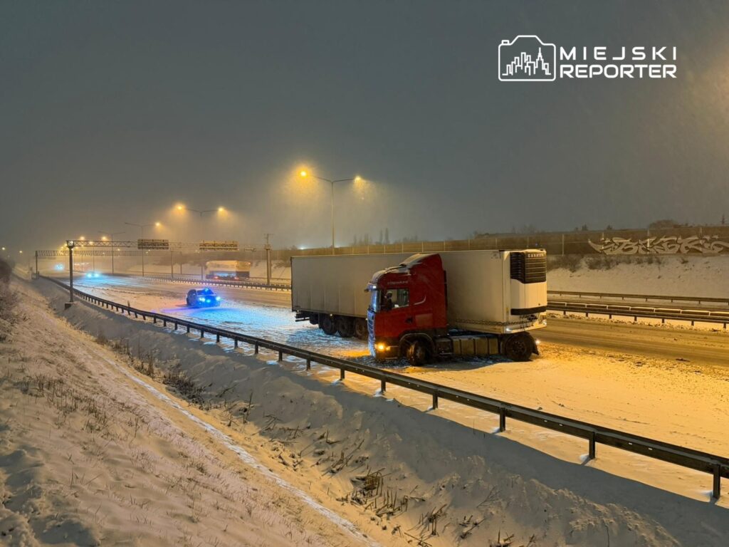 Czerwony ciężarowy pojazd utknął na zaśnieżonej drodze, obok widać radiowóz z niebieskimi światłami.