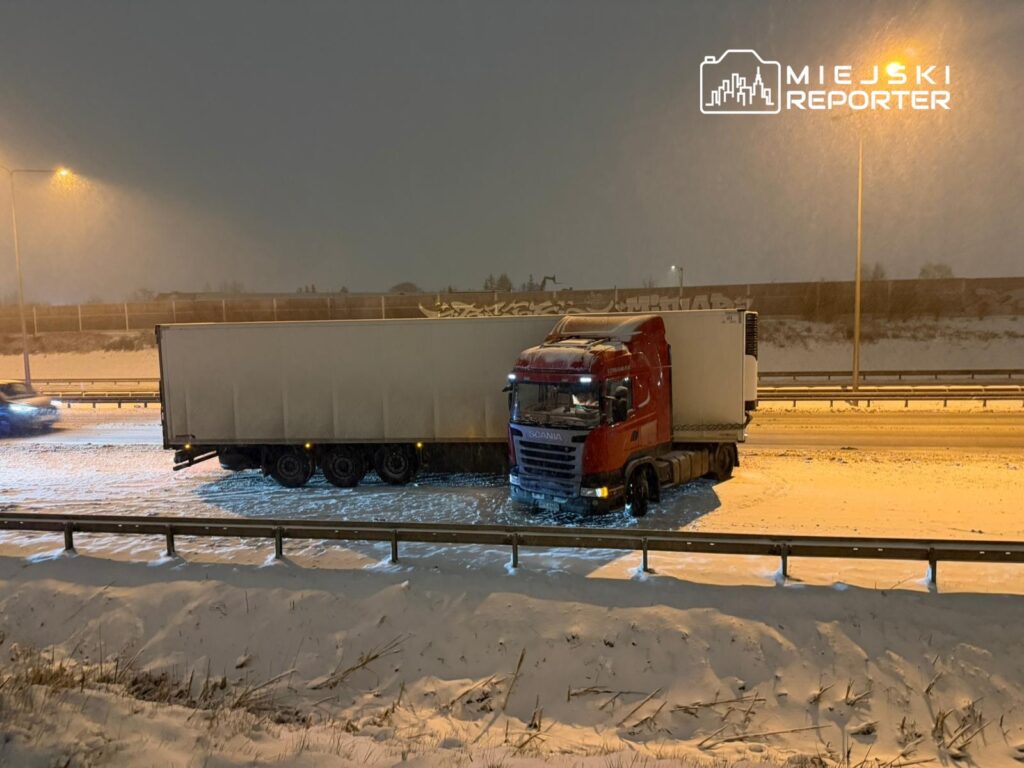 Czerwony ciężarowy samochód utknął na zaśnieżonej autostradzie, obok widoczna jest płot i padający śnieg.
