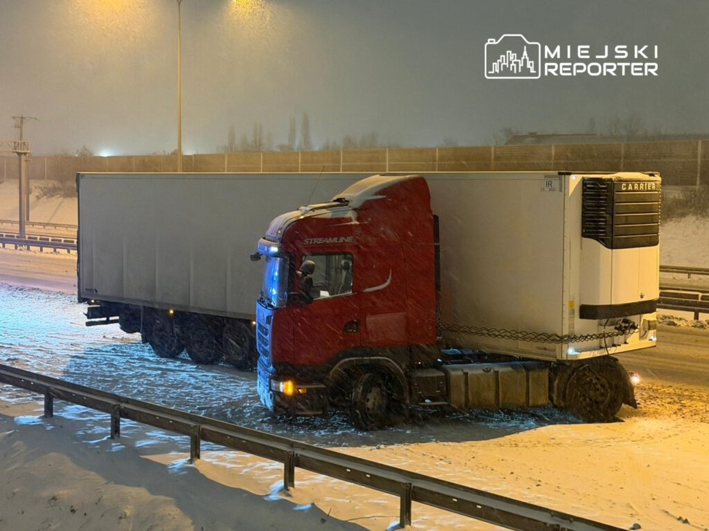 Czerwony ciągnik siodłowy z naczepą stoi na zaśnieżonej drodze pod latarnią w trakcie burzy śnieżnej.