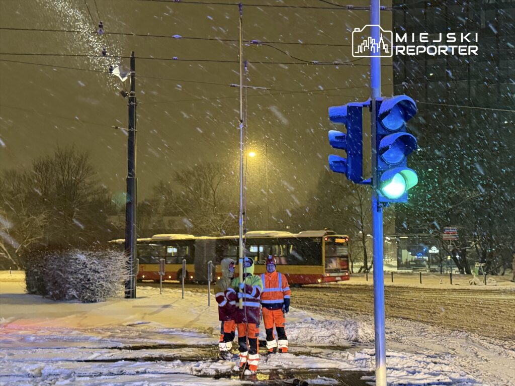 Trzech pracowników w odblaskowych kamizelkach obserwuje ruch uliczny w trakcie intensywnej śnieżycy.