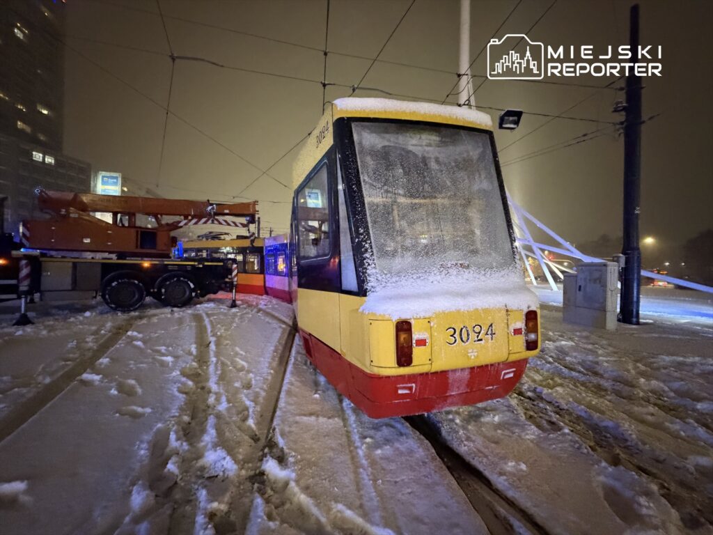 Tramwaj pokryty śniegiem stoi na zaśnieżonym torowisku, obok uniesiona dźwig z czerwonym ramieniem.