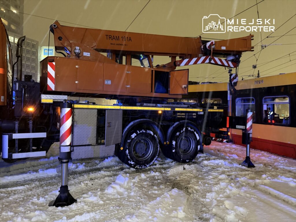 Pomarańczowy dźwig tramwajowy stojący na zaśnieżonej ulicy, obok tramwaju. W tle oświetlone budynki.