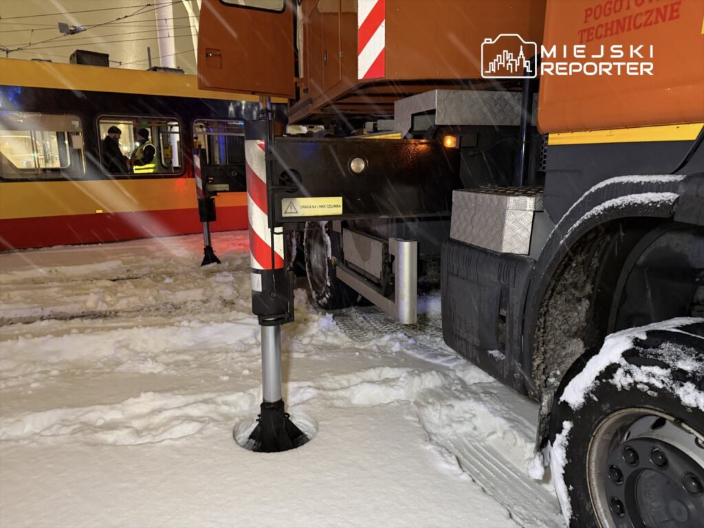 Pojazd techniczny z podniesionymi podpórkami stoi na zaśnieżonej ulicy obok tramwaju, w czasie opadów śniegu.