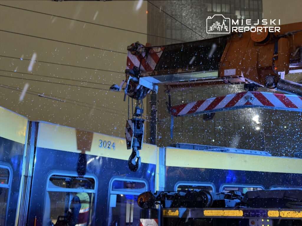 Dźwig z wciągarką pracujący przy tramwaju w trakcie opadów śniegu, otoczenie oświetlone niebieskimi światłami.
