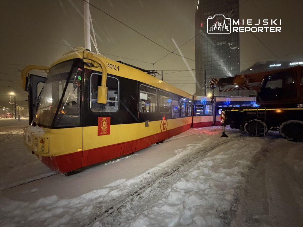 Tramwaj w kolorze żółto-czerwonym utknął w śniegu, obok stoi pojazd ratunkowy z włączonymi światłami.