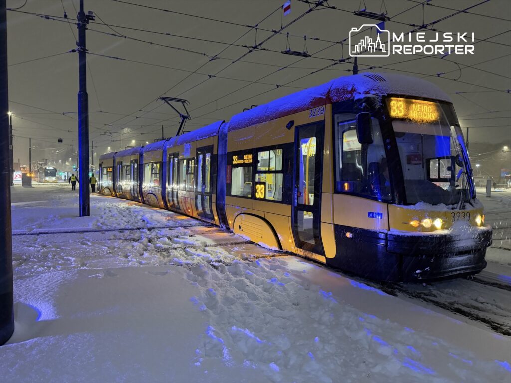 Tramwaj linii 33 zatrzymany na przystanku w zasypanym śniegiem otoczeniu, oświetlony niebieskimi i pomarańczowymi światłami.