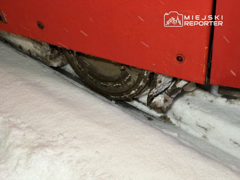 Czerwony pojazd na śnieżnej nawierzchni, z widocznymi kołami i śladami po torach.