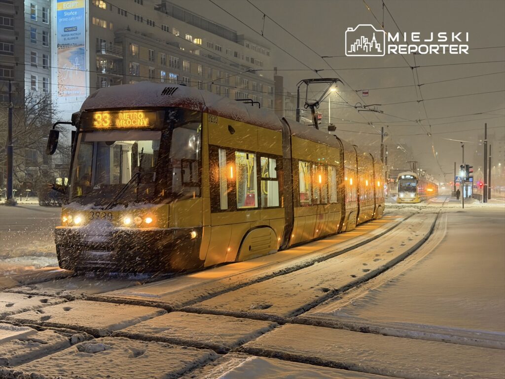 Tramwaj w zimowej scenerii zatrzymany na przystanku, z widocznym wyświetlaczem "33 METRO MOCINY" w zasypanym śniegiem otoczeniu.