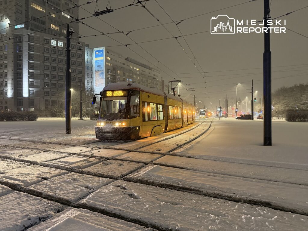 Tramwaj o numerze 33 zatrzymany na przystanku w zimowej scenerii, pokryty śniegiem, otoczony oświetlonymi budynkami.