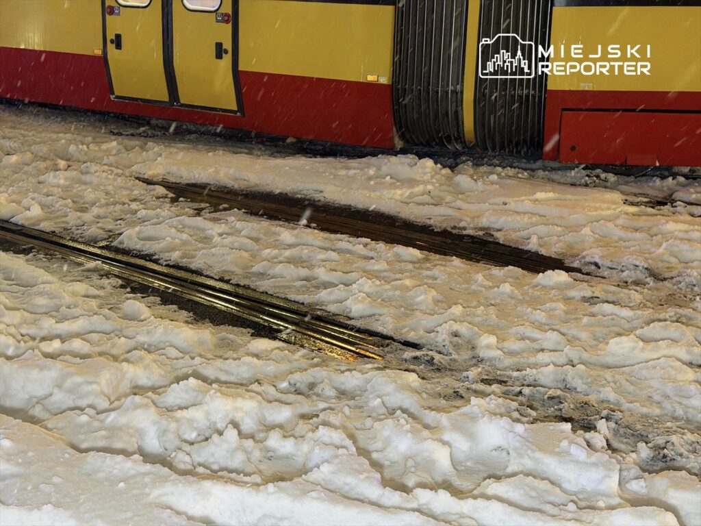 Śnieg pokrywa tory tramwajowe obok żółto-czerwonego tramwaju w zimowej scenerii.