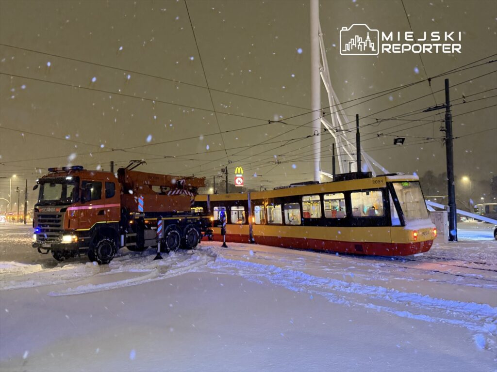 Czerwony samochód ciężarowy z podnośnikiem stoi na zaśnieżonym skrzyżowaniu obok tramwaju w zimowej scenerii.