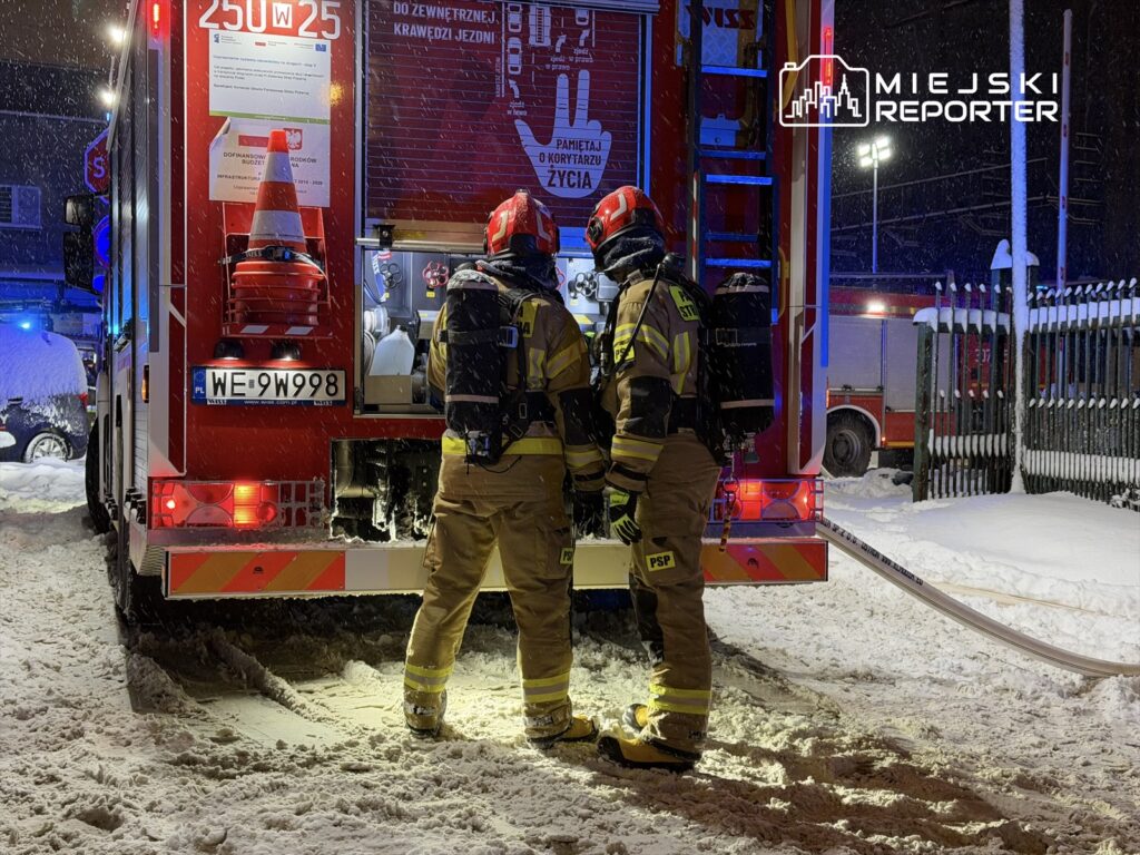 Dwaj strażacy w mundurach rozmawiają przy tylnej części wozu strażackiego na zaśnieżonej ulicy nocą.