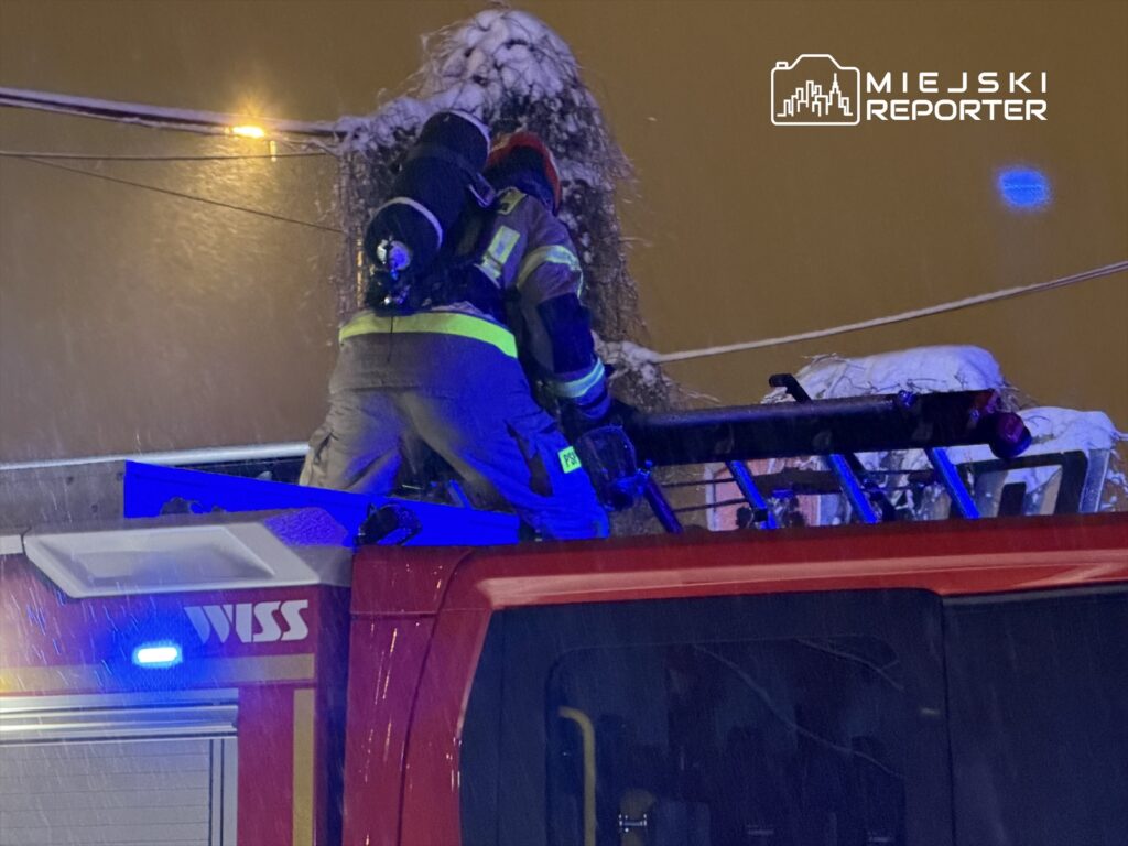 Strażak w specjalistycznym ubraniu usuwa śnieg z dachu wozu strażackiego podczas intensywnej śnieżycy.