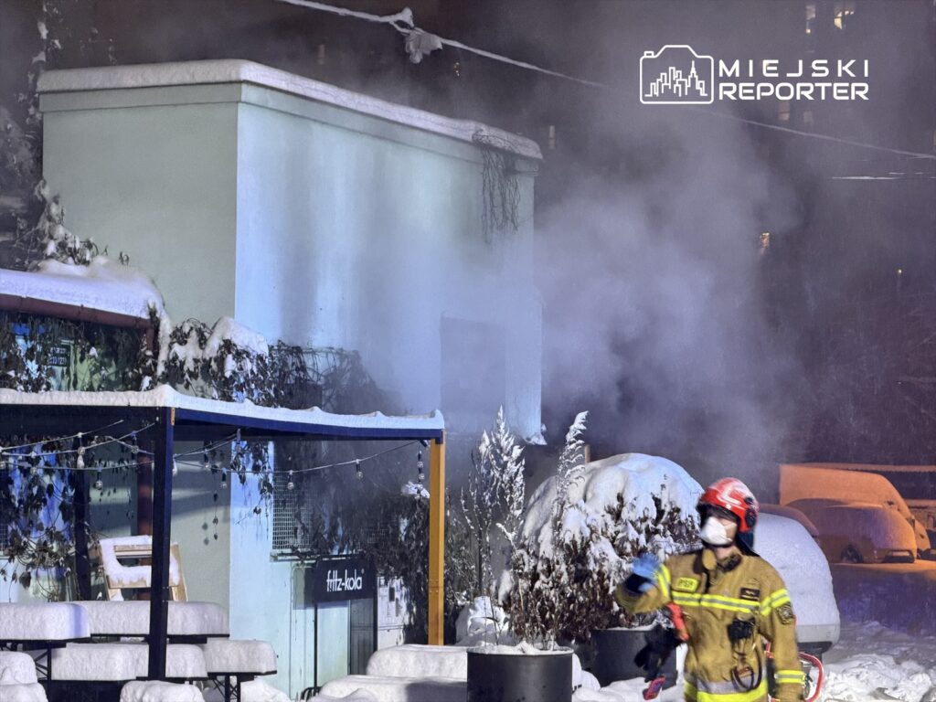Strażak w mundurze gaśniczym podchodzi do budynku z którego wydobywa się dym, otoczenie pokryte śniegiem.