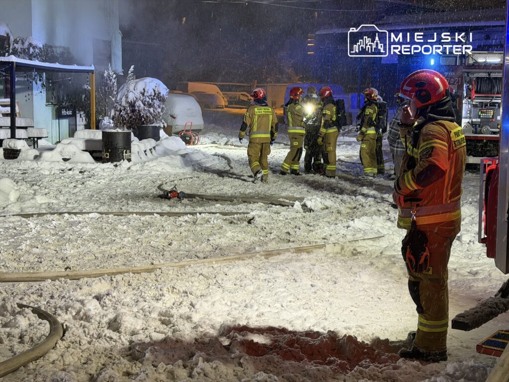 Grupa strażaków w odzieży ochronnej pracuje w zaśnieżonym terenie, gasząc pożar przy budynku.