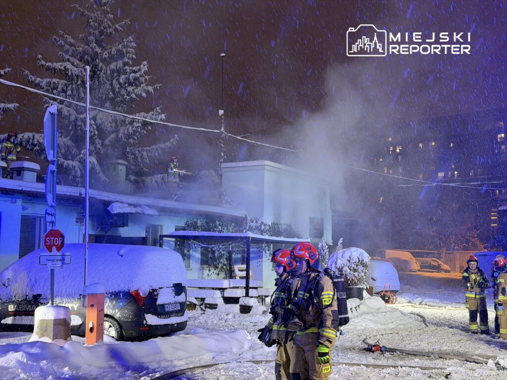 Strażacy w specjalnych strojach gaszą pożar na dachu budynku w trakcie intensywnego opadu śniegu w nocy.