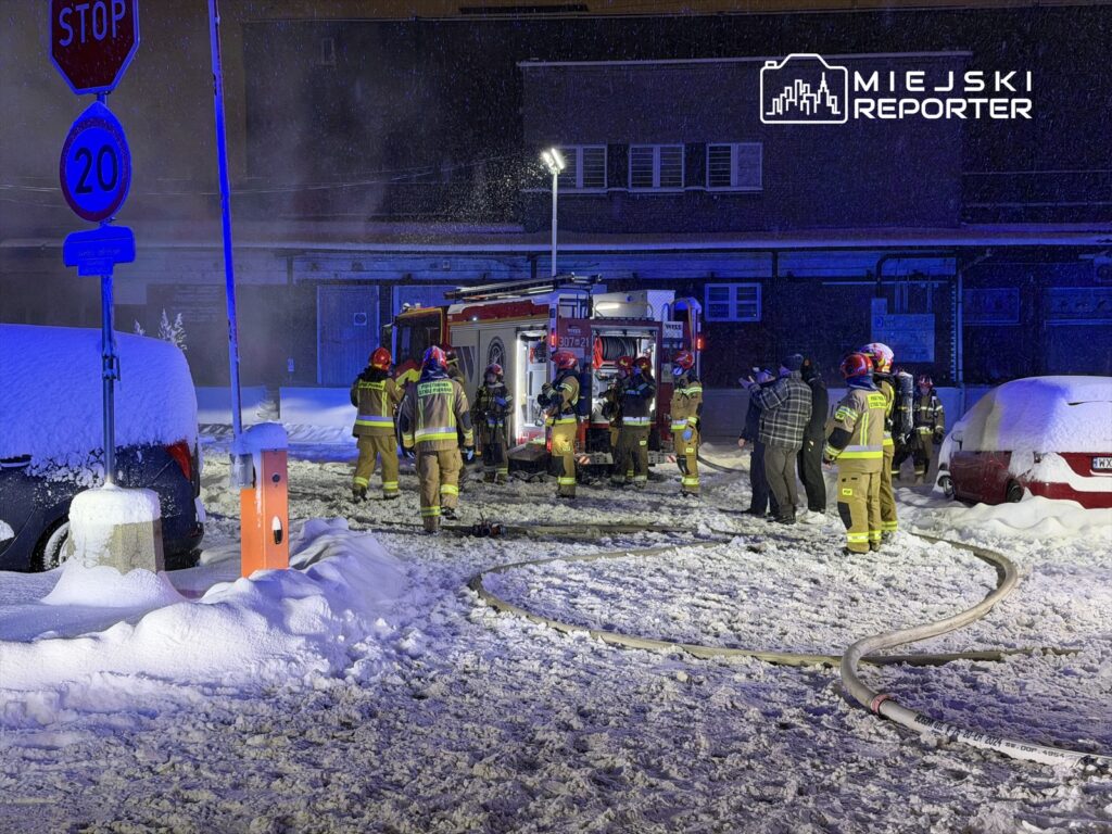 Grupa strażaków w specjalnych strojach rozmawia obok wozu strażackiego na zaśnieżonej ulicy nocą.