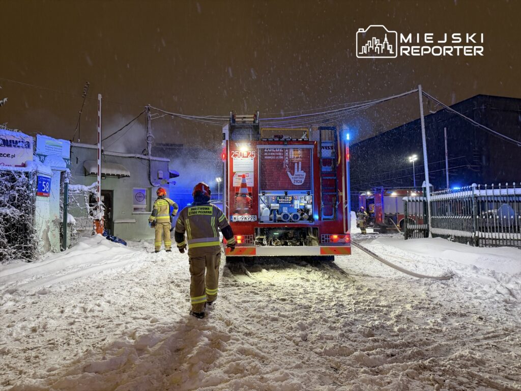 Strażacy w zimowych mundurach pracują przy wozie strażackim na zaśnieżonym terenie, w tle widać dym.