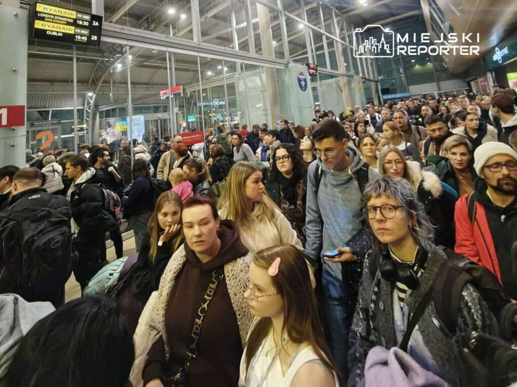 Tłum ludzi czekających w terminalu lotniska, z tablicą odlotów w tle. Wszyscy mają bagaże i wyglądają na zniecierpliwionych.