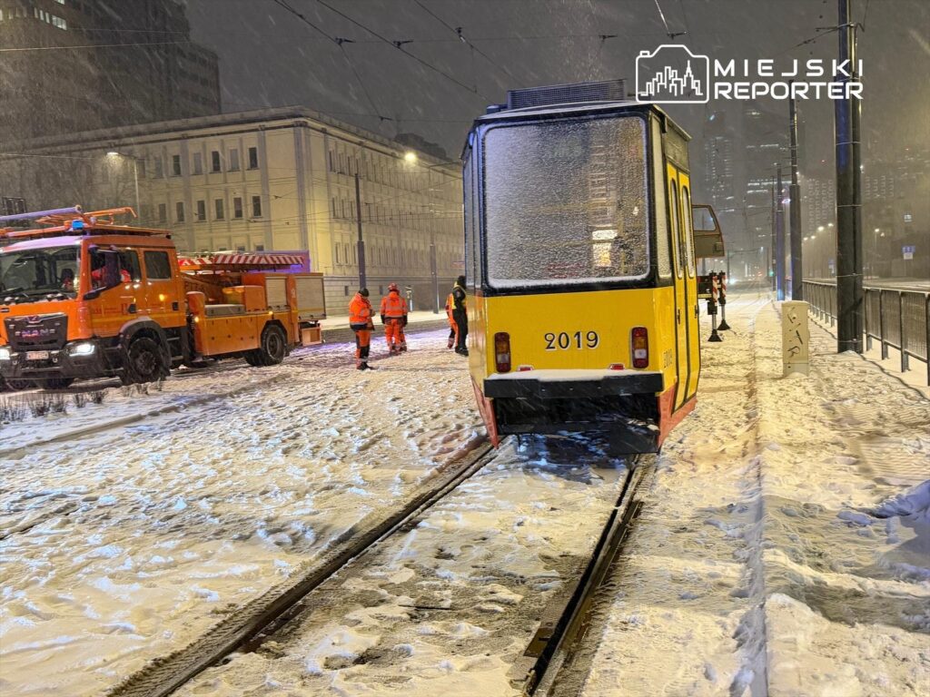 Zimowy krajobraz z tramwajem na torach, obok pomarańczowy wóz strażacki oraz pracownicy w odzieży roboczej.