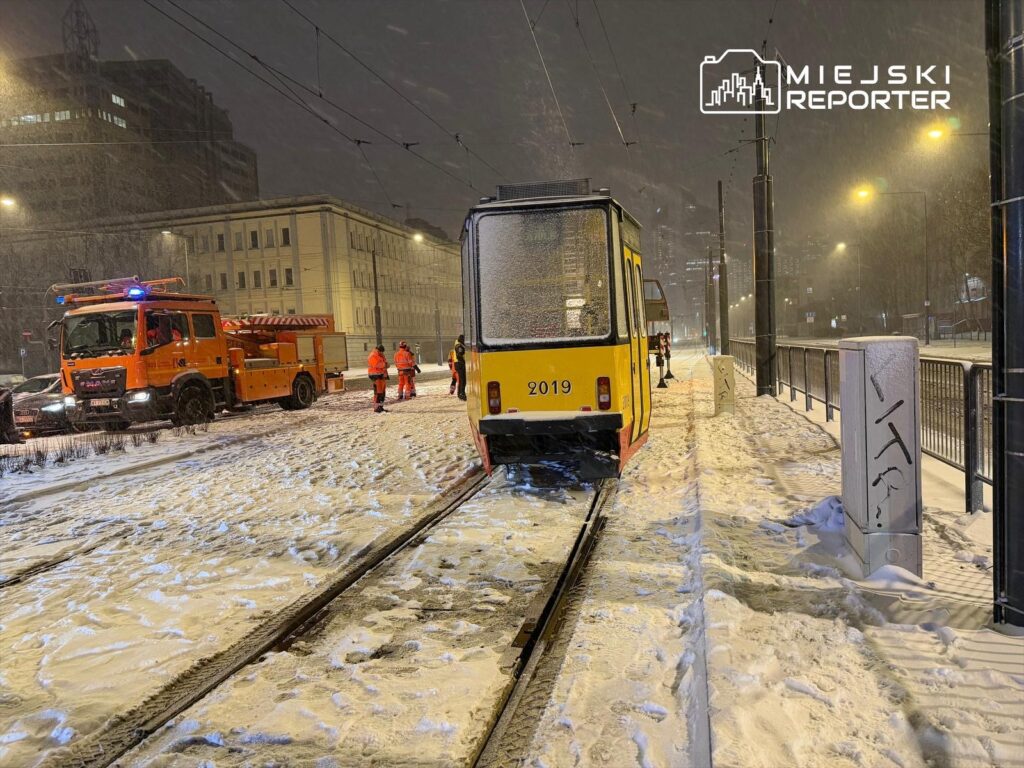 Zespół ratunkowy w pomarańczowych strojach pracuje przy tramwaju na zaśnieżonej ulicy w nocy.