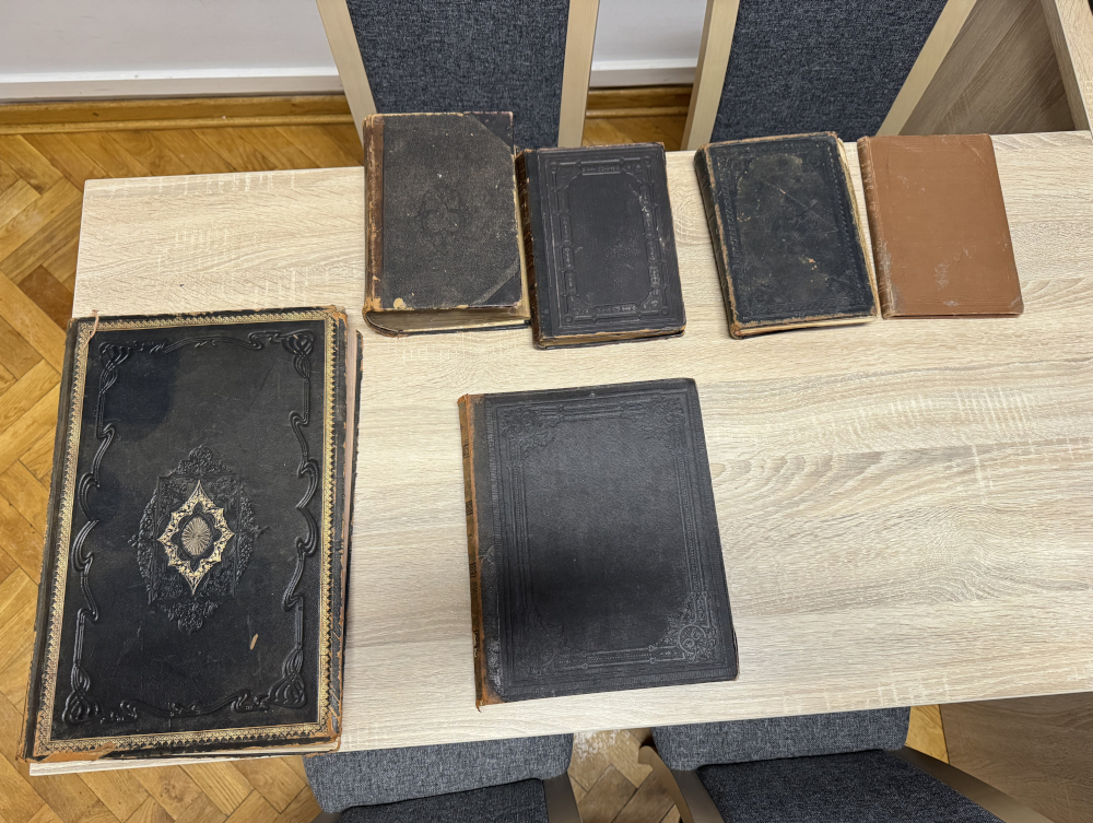 Na drewnianym stole leży sześć starych książek, w tym dwie z bogato zdobionymi okładkami i cztery o prostszej formie.