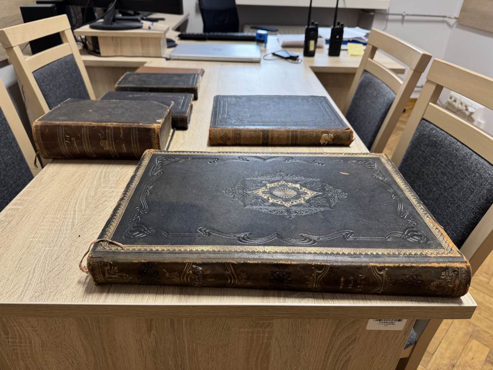 Stare, oprawione w skórę księgi leżą na drewnianym biurku w biurze, obok nowoczesnego sprzętu komputerowego.