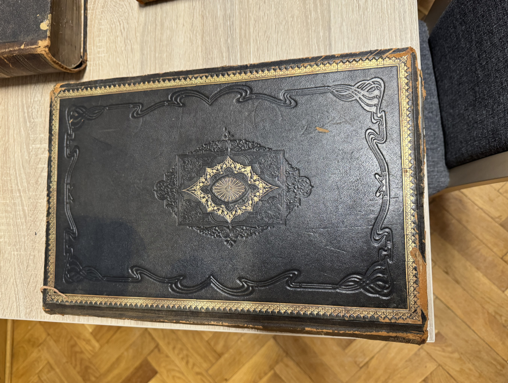 Czarny, skórzany album z ozdobnym tłoczeniem i złotymi detalami leży na drewnianym stole.