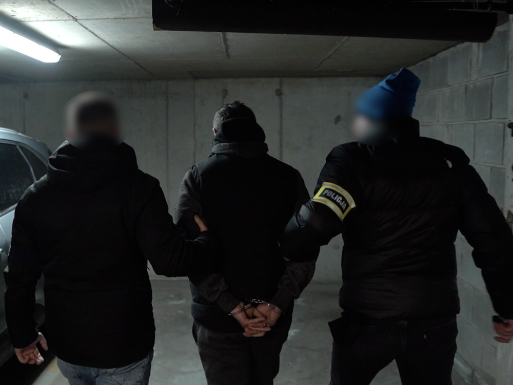 Mężczyzna z rękami skuty w kajdankach prowadzony przez dwóch funkcjonariuszy w garażu.