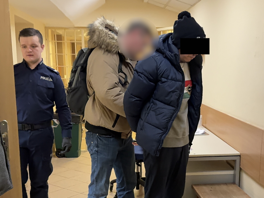 Mężczyzna w granatowej koszuli policyjnej prowadzi zatrzymanego mężczyznę w kurtce do pomieszczenia.