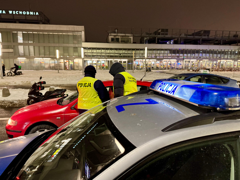 Dwaj policjanci w żółtych kamizelkach kontrolują samochód osobowy na parkingu przy dworcu.