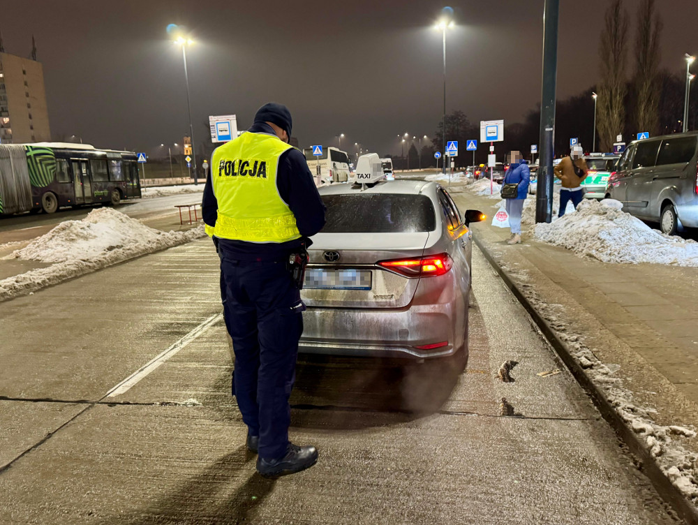 Policjant w kamizelce odblaskowej kontroluje taksówkę na oświetlonym parkingu w zimowej scenerii.