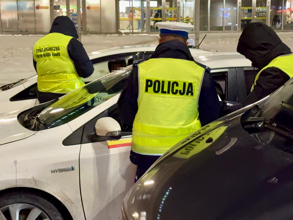 Funkcjonariusze policji w kamizelkach odblaskowych kontrolują samochód na parkingu w nocy.