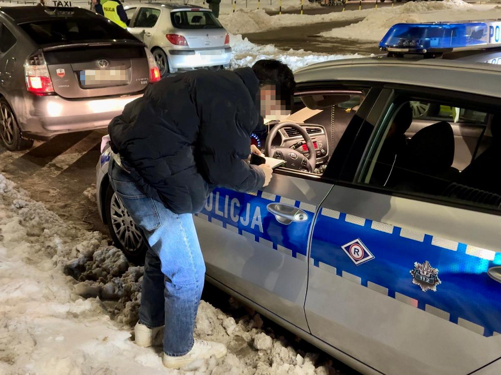 Mężczyzna w kurtce podchodzi do policyjnego radiowozu zaparkowanego na zaśnieżonej ulicy.
