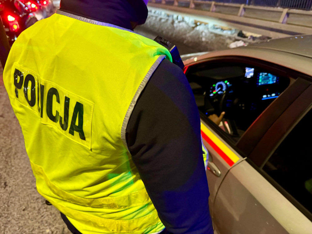 Policjant w kamizelce odblaskowej kontroluje kierowcę w samochodzie na ulicy nocą.