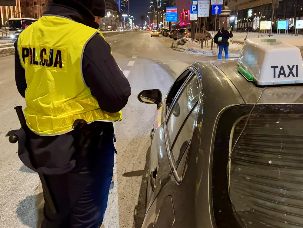 Policjant w żółtej kamizelce rozmawia z kierowcą taksówki na oświetlonej ulicy w nocy.