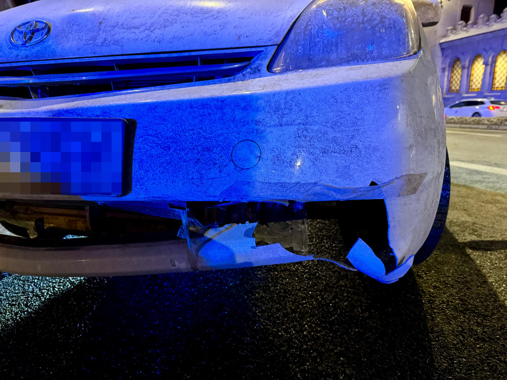 Uszkodzony przód białego samochodu osobowego na ciemnej ulicy, oświetlony niebieskim światłem.